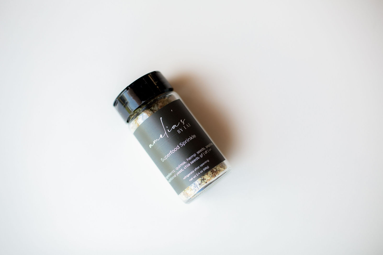 Amelia's Superfood Sprinkle in a black-capped glass shaker jar lying on a white surface