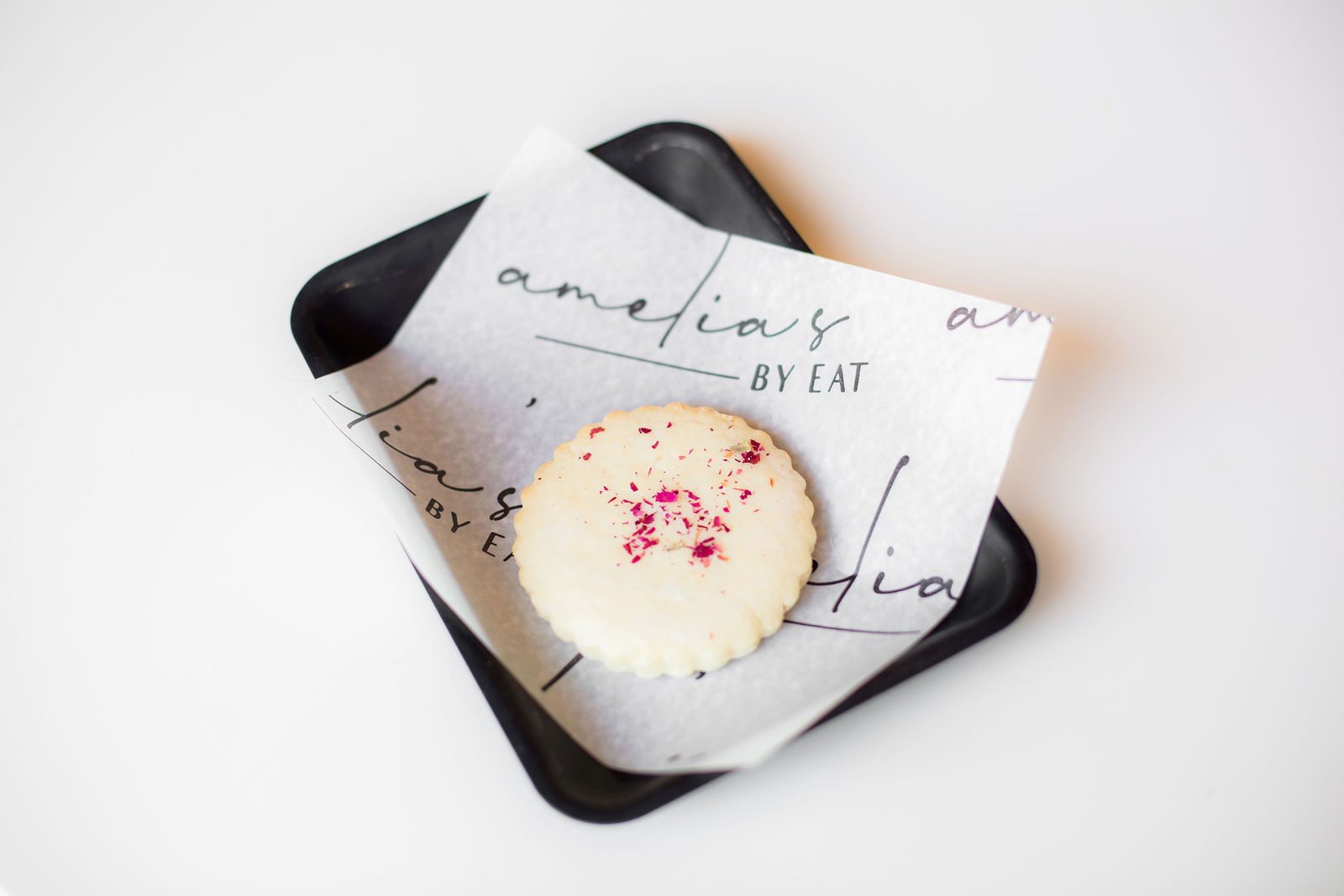 Glazed scalloped sugar cookie with dried rose petals on Amelia's branded paper and black tray