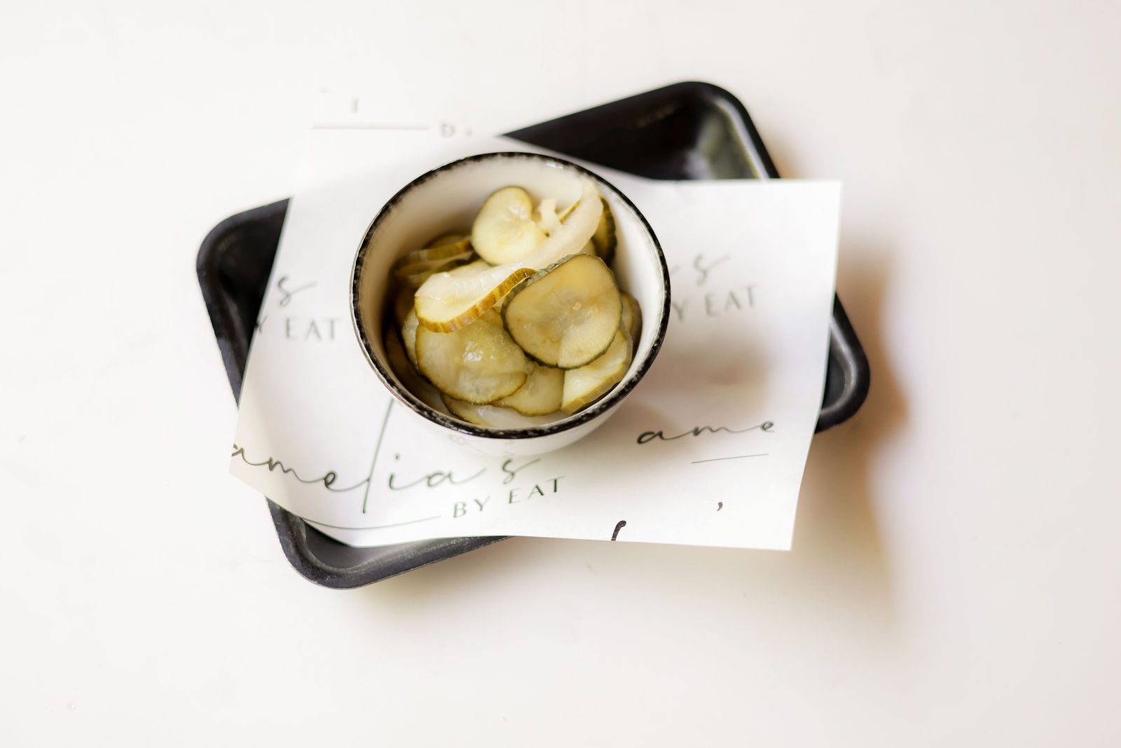 Bowl of sliced housemade pickles on Amelia's by EAT branded paper and a black tray