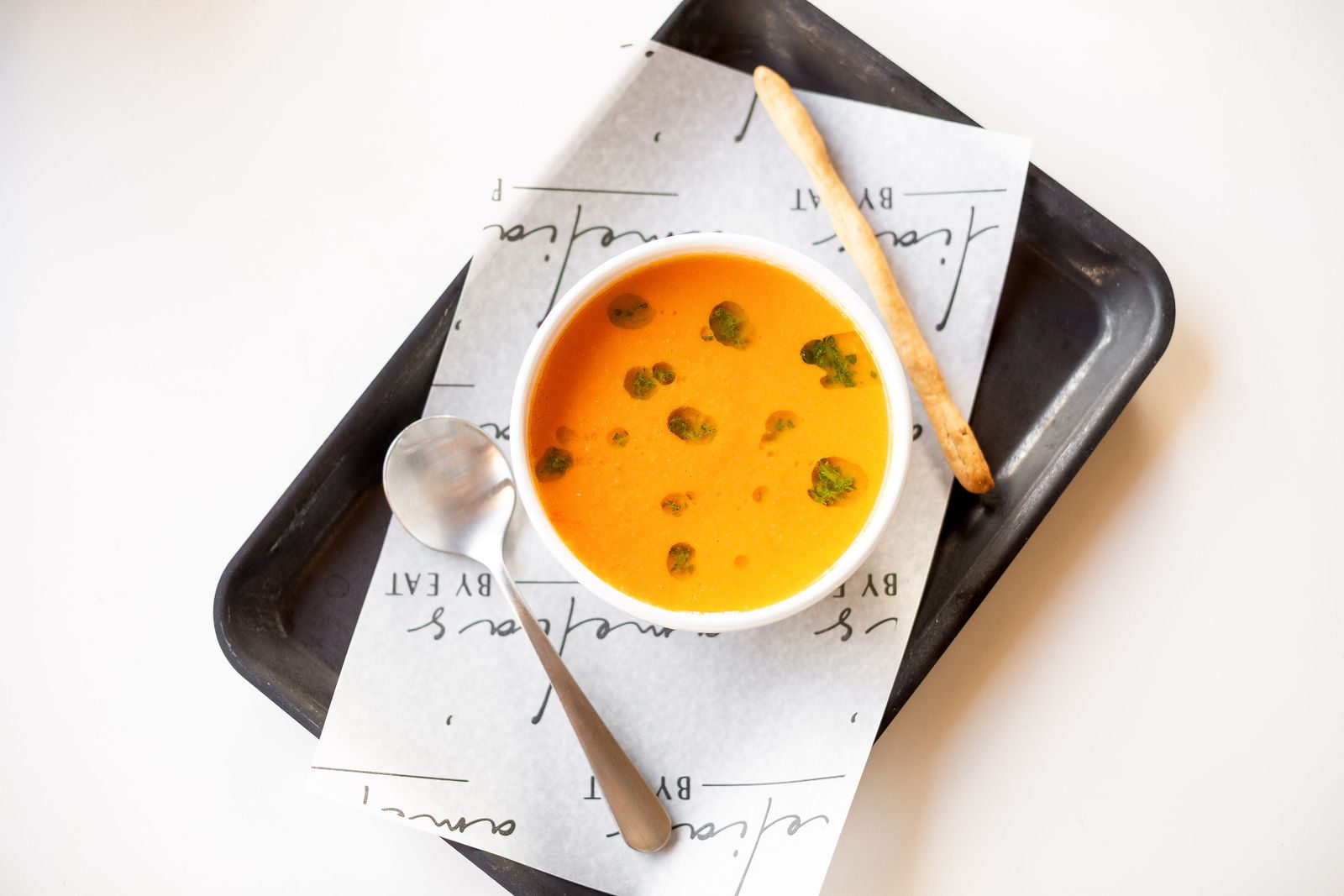 Bowl of tomato soup with green herb oil, breadstick and spoon on Amelia's paper